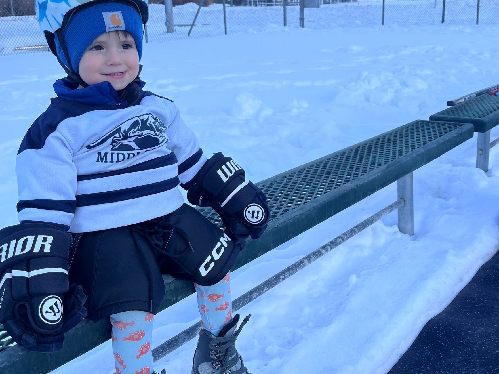 Child in skates and helmet smiling next to the outdoor ice rink. 